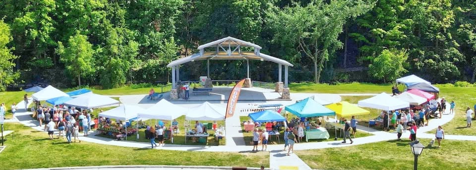 Aerial view of an outdoor farmers market set around a pavilion in a park. Multiple white and colorful vendor tents line a circular walkway, with shoppers browsing booths and gathering throughout the market. Trees and wooded hillsides surround the venue on a sunny summer day.