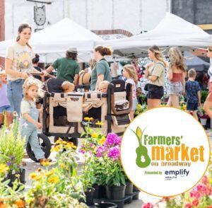 Families and shoppers browse vendor tents at the Farmers Market on Broadway in downtown Green Bay. Flower and plant vendors line the foreground, while adults and children walk through the market. A logo for Farmers Market on Broadway appears in the lower right corner, and text at the top notes the market runs Wednesdays from May through September.